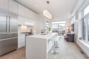 Modern white kitchen with a large island and bar stools, opening to a living area with natural light.