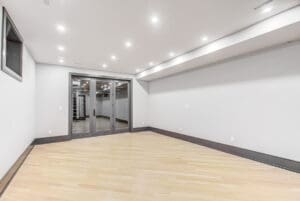 Empty modern basement room with light wood flooring and mirrored wall.