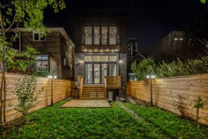 A modern house with a wooden deck and fenced backyard at night.