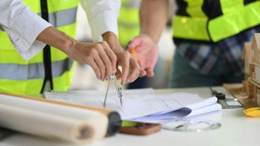 Hands using a compass to draw on architectural plans at a construction site.